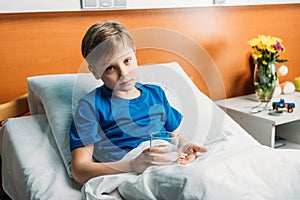 Upset boy holding glass of water and medicines in hospital bed