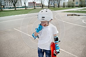 Upset boy skateboarder in protective gear frustratedly wipes tears from his eyes and holds a skateboard in one hand while standing