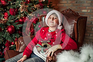 Upset boy in a vintage chair near a Christmas tree with red balls, Boy in a Santa Claus hat waiting for gifts in a deer sweater in