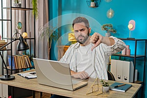 Upset business man freelancer working on laptop showing thumbs down sign disapproval dissatisfied