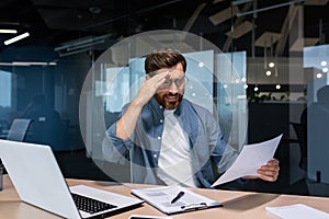 Upset businessman behind paper work inside modern office, mature man with beard reading financial reports and account