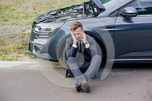 Upset businessman use phone sitting on road near the broken car opened the hood help repair