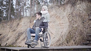 Upset Caucasian boy sitting in wheelchair with mother or invalid tender. Sad crippled man spending autumn day outdoors