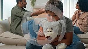 Upset Caucasian little boy preschool son hug teddy toy while African American woman mother and man father in anger