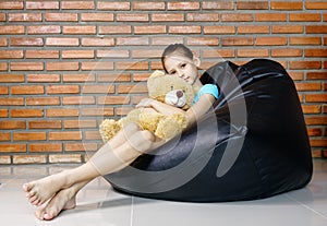 Upset caucasian teen girl sitting in black bean bag chair holding soft teddy bear toy against brick wall. Casual outfit. Child