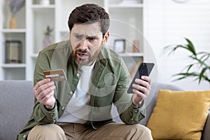 Upset cheated man sitting on couch in living room at home, holding bank credit card and phone, rejected money transfer