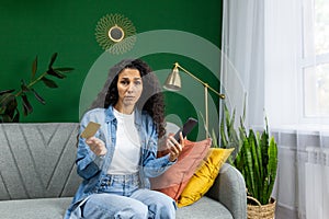 Upset cheated woman looking at camera, holding phone and bank credit card, sitting on sofa at home in living room