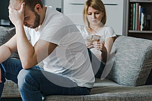 Upset couple sitting at the sofa. Woman counts last money while her stressed husband holding his head with hands.