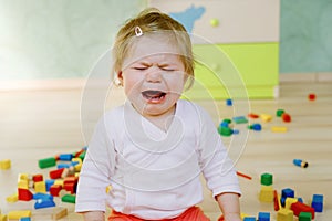 Upset crying baby girl with educational toys. Sad tired or hungry alone healthy child sitting near colorful different