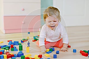 Upset crying baby girl with educational toys. Sad tired or hungry alone healthy child sitting near colorful different