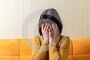 Upset crying middle aged woman suffering from stress covering her face with her hands on an orange sofa