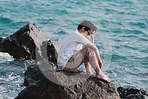 Upset depressed Asian man with hands on face feeling bad at rock of natural seashore