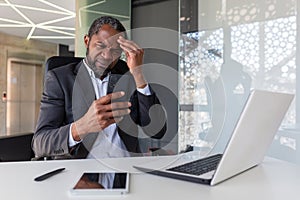 Upset and disappointed man inside office holding phone, businessman reading bad news at workplace inside office, african