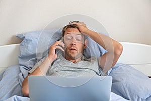 Upset disappointed man with mobile phone disputing looking at laptop while laying in a bed at home and solving online