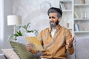 Upset and disappointed man sitting on sofa at home, hispanic received mail message letter with bad news, sad depressed