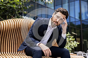 Upset and disappointed young businessman man sits on a bench near an office and court building and holds his head