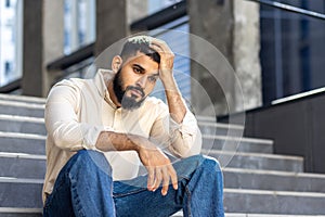 Upset and disappointed young Indian man sitting on the steps of an office building and holding his hand above his head