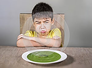 Upset and disgusted latin kid sitting on table refusing to eat spinach thick soup looking unhappy rejecting vegetarian food in