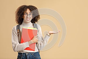 Upset doubt african american woman in glasses with backpack holds notepads and shows hand to blank space