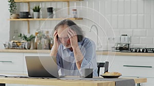 upset elderly man near a laptop in the kitchen. problems with online service, bad news, big bills