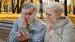 Upset elderly married couple sit on bench in autumn park read bad news on smartphone worried old people look at screen