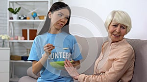 Upset elderly woman refusing to eat oatmeal offered by volunteer, nursing home