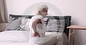 Upset elderly woman sitting on bed rubbing back feeling backache