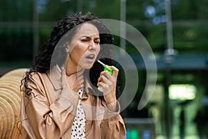 Upset female holding small green bottle and spraying throat spray into mouth to alleviating pain outdoors. Lady wearing