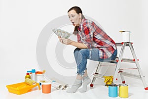 Upset female holds bundle of dollars, cash money, sits on ladder with instruments for renovation apartment isolated on