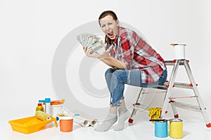 Upset female holds bundle of dollars, cash money, sits on ladder with instruments for renovation apartment isolated on