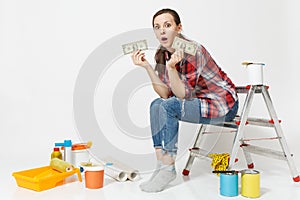 Upset female holds tight of dollars, cash few money, sits on ladder with instruments for renovation apartment isolated