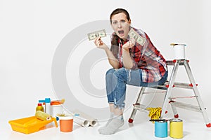 Upset female holds tight of dollars, cash few money, sits on ladder with instruments for renovation apartment isolated