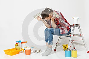 Upset female holds tight of dollars, cash few money, sits on ladder with instruments for renovation apartment isolated