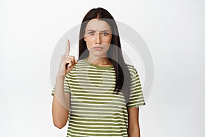 Upset frowning girl pointing finger up, look jealous or disappointed, standing in t-shirt over white background