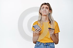 Upset girl holding smartphone and credit card, concept of woman lacking money, cant buy something, standing over white