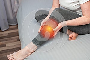 Upset girl looking at camera while sitting on bed and suffering from pain in leg. Tendon knee joint problems on woman leg