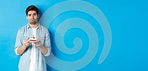 Upset and gloomy man holding birthday cake, sulking sad, standing against blue background