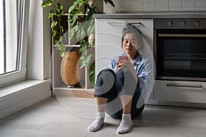 Upset gloomy young Asian woman looking at mobile phone screen feeling sad while sitting on floor