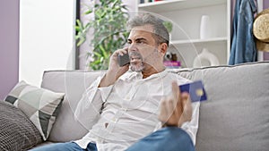 Upset hispanic man with grey hair in a problematic phone talk, holding a credit card at home, expressing worry indoors