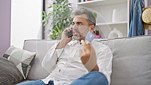 Upset hispanic man with grey hair in a problematic phone talk, holding a credit card at home, expressing worry indoors