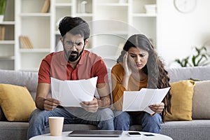 Upset indian couple checking bills, sitting on couch, reading documents