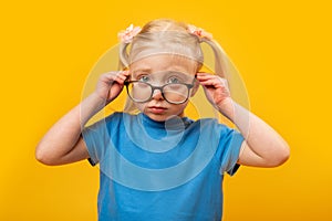 Upset little blond girl adjusts her glasses and looking straight. Portrait of girl in blue T-shirt with glasses on yellow
