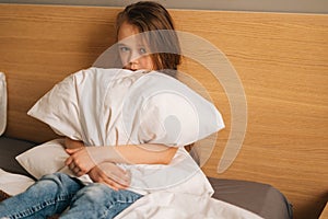 Upset little girl crying sitting alone in fetal position and rocking on bed hugging big pillow and looking at camera.