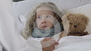 Upset little girl lying in bed, having high temperature and looking at physician
