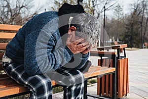 Upset lonely homeless elderly old Caucasian man with depression holds head in hands while sitting on park bench in