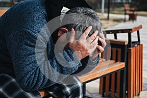 Upset lonely homeless elderly old Caucasian man with depression holds head in hands while sitting on park bench in