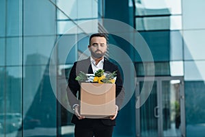 Upset male office worker holding box with things and leaving office building, free space