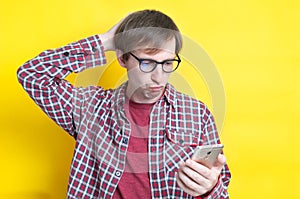 Upset man in checkered shirt with hand on back of head and in glasses looking at smartphone