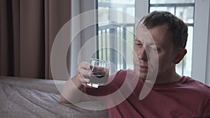 Upset man drinking alcohol while sitting on sofa, window background