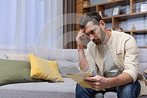 Upset man at home sitting on sofa reading letter with bad news notification in envelope opening, unhappy in living room